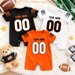 Könnte beinhalten: Drei kurzärmelige Baby-Strampler in Schwarz, Weiß und Orange, jeweils mit dem Text TEAM NAME und der Zahl 00 in fetter Schrift. Die Ärmel haben schwarze Tigerstreifenmuster. Die Strampler verfügen über Druckknöpfe aus Metall.