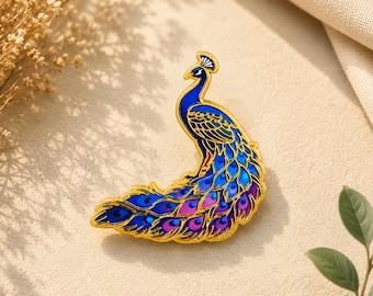 Könnte beinhalten: Eine goldfarbene Emaille-Anstecknadel in Form eines Pfaus. Der Pfau hat einen leuchtend blauen Hals und Kopf mit einem Kamm sowie einen langen, geschwungenen Schwanz mit komplizierten Federmustern in Blau-, Violett- und Goldtönen.