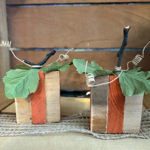Pode incluir: Duas abóboras decorativas feitas de blocos de madeira retangulares em tons de madeira natural, laranja e cinza. Cada abóbora apresenta um caule de galho marrom escuro, folhas de tecido verde e gavinhas de arame em espiral no topo.