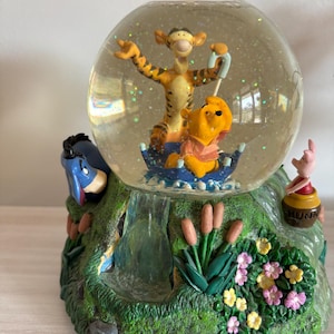May include: A snow globe featuring Tigger and Winnie the Pooh inside a blue boat. The base is a green landscape with a waterfall, cattails, and flowers. Eeyore is on the left side, and Piglet sits on a honey pot labeled HUNNY on the right.