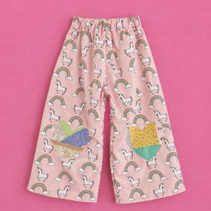 May include: Light pink wide-leg pants featuring an all-over print of white unicorns, colorful rainbows, and clouds. Each leg features a large, heart-shaped patch constructed from various floral and solid-colored fabric pieces in pastel tones.