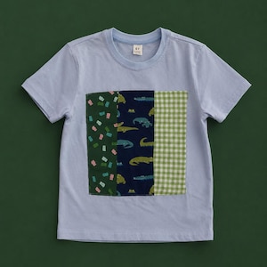 May include: A light blue short-sleeve t-shirt featuring a central rectangular patch made of three vertical fabric panels. The panels display a dark green gummy bear print, a navy blue alligator pattern, and a light green and white gingham check.