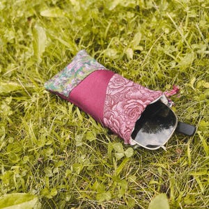 May include: A fabric glasses case featuring a patchwork design with a green floral pattern, a solid magenta center panel, and a pink rose print section, containing a pair of black-framed sunglasses partially visible at the open end of the pouch.