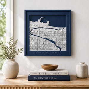 May include: A square, dark blue framed wall art piece featuring a three-dimensional, white relief map of Traverse City, Michigan. The map displays detailed street grids and a waterfront area, with the text TRAVERSE CITY, MICHIGAN embossed below.