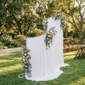 Puede incluir: Un telón de fondo de tela blanca con dos paneles curvos de diferentes alturas, cubiertos con material suave y fluido. La estructura está adornada con tres arreglos florales de rosas blancas y color melocotón mezcladas con hojas de eucalipto.