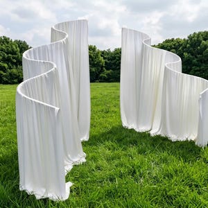 Può includere: Due alte strutture autoportanti in tessuto bianco con un design verticale ondulato a forma di onda. Il materiale bianco è drappeggiato in pieghe morbide e fluide, creando una forma curva e serpentina che si erge verticalmente.