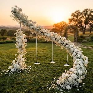 Peut inclure: Une structure en métal incurvée décorée de fleurs blanches et de gypsophile. L'arrangement floral suit la forme de l'arche, soutenu par trois fins poteaux verticaux blancs avec des bases circulaires posés sur une pelouse herbeuse.