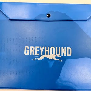 May include: A bright blue plastic document folder with a flap closure and a single black snap button. The word GREYHOUND is printed in white capital letters above a white silhouette graphic of a running greyhound dog on the front surface.