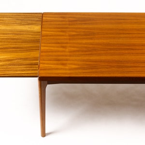 May include: A wooden dining table with a leaf extension. The table is made of light brown wood with a natural grain pattern. The leaf is attached to the table with hinges and can be folded down when not in use.
