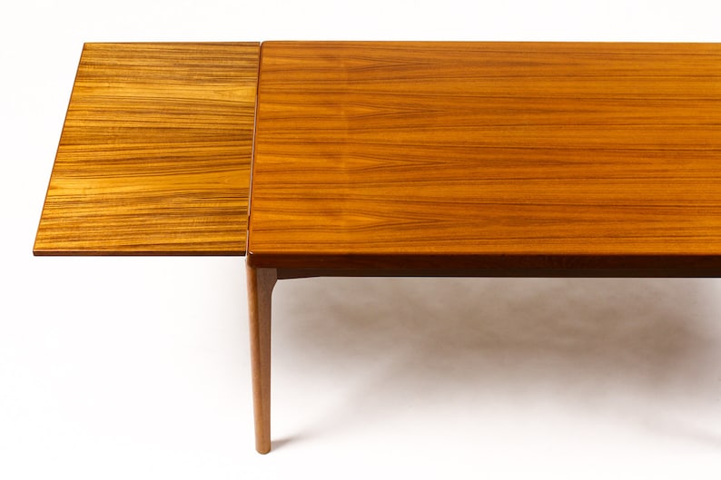 May include: A wooden dining table with a leaf extension. The table is made of light brown wood with a natural grain pattern. The leaf is attached to the table with hinges and can be folded down when not in use.