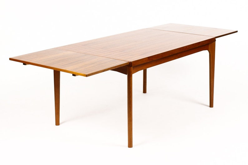 May include: A wooden extendable dining table with a light brown finish. The table has a rectangular top and four legs. The table is shown with one leaf extended.
