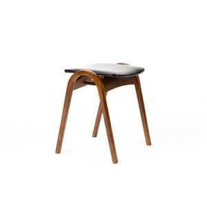 May include: A mid-century modern stool with a black leather seat and curved wooden frame. The stool has three angled legs and a curved back support. The wood has a warm, brown tone, and the design is minimalist.