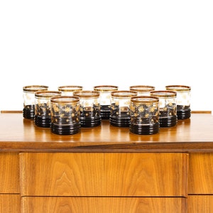 May include: A set of 12 clear glass tumblers with gold accents and black bottoms, arranged on the top of a wooden dresser with two drawers.