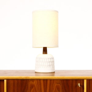 May include: A white ceramic table lamp with a textured base and a white fabric lampshade. The lamp is sitting on a wooden table.