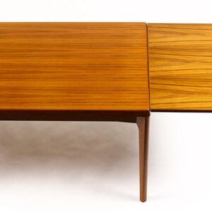 May include: A wooden extendable dining table with a light brown finish. The table has a rectangular top and four legs. The table is shown with one leaf extended.