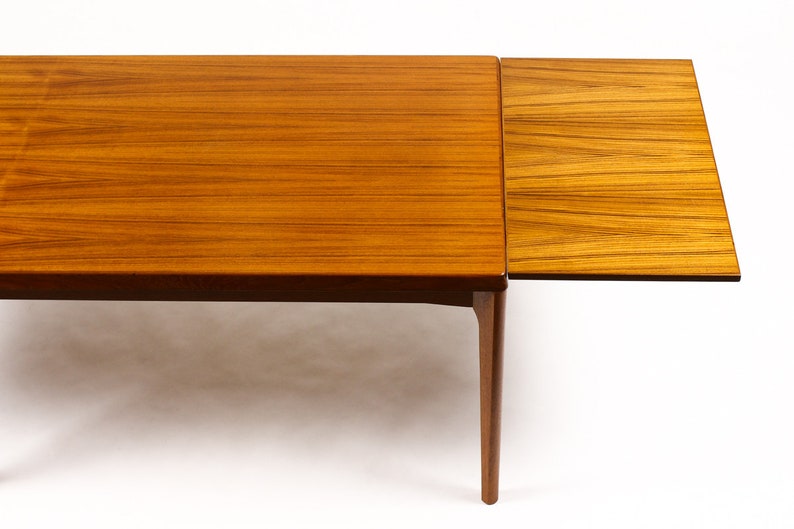 May include: A wooden extendable dining table with a light brown finish. The table has a rectangular top and four legs. The table is shown with one leaf extended.