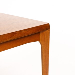 May include: Close-up of the corner of a square wooden table with a light brown finish. The table has a simple, modern design with slightly curved legs.