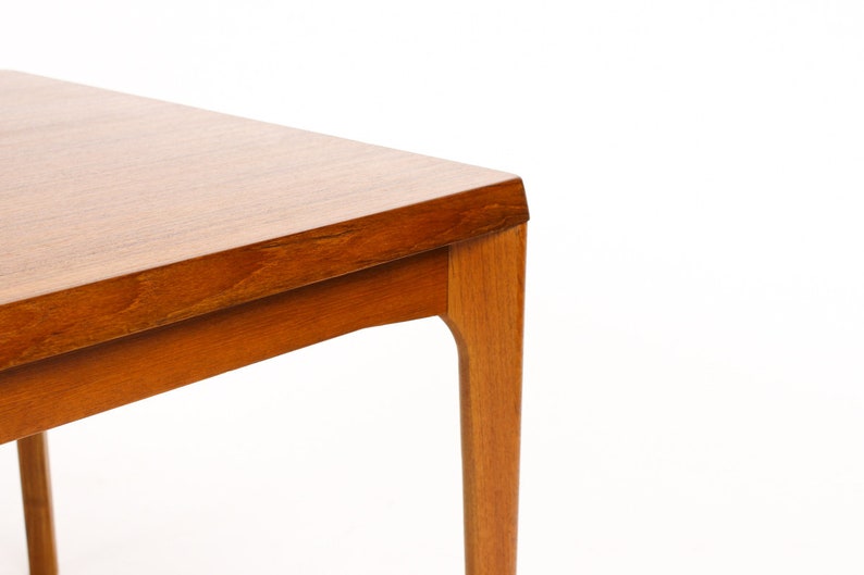 May include: Close-up of the corner of a square wooden table with a light brown finish. The table has a simple, modern design with slightly curved legs.