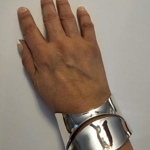 May include: A wide, polished silver-tone cuff bracelet featuring a smooth, curved, overlapping design. The metal surface is highly reflective, creating a mirror-like finish that catches light across its rounded, sculptural form.