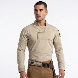 Könnte beinhalten: Ein beiges Langarmshirt mit hohem Kragen und überlappendem Vorderteil, kombiniert mit einer braunen Cargohose. Ein Utility-Gürtel mit Karomuster, einer Metallschnalle und einer dunkelbraunen Oberschenkeltasche mit Riemen wird um die Taille getragen.