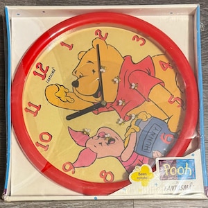 May include: A round wall clock with a bright red frame and a yellow face featuring Winnie the Pooh and Piglet. The clock has red numbers, black hands, and small bee graphics. A label on the packaging reads Pooh by Fantasma and Bees rotate!