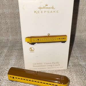 May include: A Hallmark Keepsake Lionel Union Pacific Streamliner Long Coach Christmas ornament in a white box. The ornament is a yellow and brown train car with a gold hanging loop. The box features the Hallmark logo and the text LIONEL Union Pacific Streamliner Long Coach.