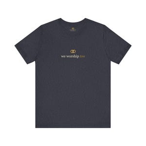 May include: A dark heather gray short-sleeve t-shirt featuring a small gold-colored graphic of two interlocking rings above the text "we worship too" in white and gold lettering, with a small brand logo printed inside the neck area.