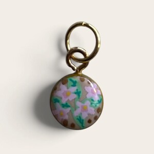May include: A small, circular gold-toned charm featuring an enamel inlay with a light brown background, pink flowers with yellow centers, and green leaves. The charm is attached to a gold-toned jump ring at the top for easy attachment to jewelry.