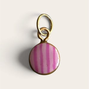 May include: A small, circular charm featuring a gold-toned metal rim and a glossy enamel inlay with vertical stripes in alternating shades of light pink and magenta. The charm is attached to a simple, circular gold-toned metal jump ring.