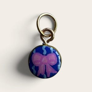 May include: A circular gold-toned metal charm featuring a glossy blue enamel surface with a hand-painted light pink bow at the center, accented by small light blue wavy lines. The charm is attached to a gold-toned jump ring for easy attachment.