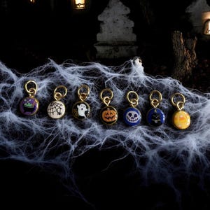 May include: A set of seven circular metal charms with gold-toned jump rings, each featuring a different Halloween-themed enamel design including a purple cauldron, spider web, white ghost, orange pumpkin, skull, black cat, and yellow candy corn.