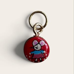 May include: A circular metal charm with a glossy red enamel finish featuring a hand-drawn style illustration of a mouse wearing a blue top and pink skirt. The charm is attached to a gold-toned metal jump ring at the top for easy attachment.