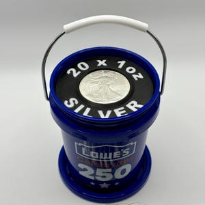 May include: A blue plastic bucket with a white handle, featuring the text Lowe's Celebrates 250 on the side. The top is covered by a black circular insert with the white text 20 x 1oz SILVER, centered around a single silver coin.