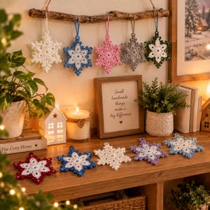 May include: A collection of crocheted snowflake ornaments in various colors including white, blue, pink, gray, green, red, and purple. Five snowflakes hang from a wooden branch, while five others are arranged on a wooden surface.