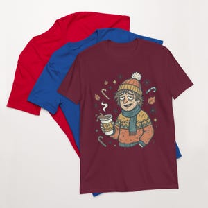 Può includere: Una pila di tre magliette a maniche corte nei colori rosso, blu e bordeaux. La maglietta bordeaux in alto presenta un'illustrazione grafica di una persona con berretto e sciarpa, che tiene in mano una tazza di caffè con la scritta Q4 FUEL.