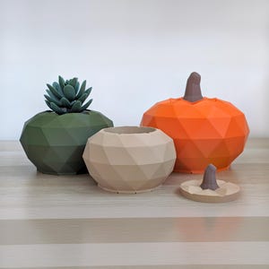 May include: Three geometric containers with faceted surfaces. One dark olive green container holds a succulent, one beige container is open, and one bright orange container has a brown stem-shaped lid, with a separate matching beige lid nearby.