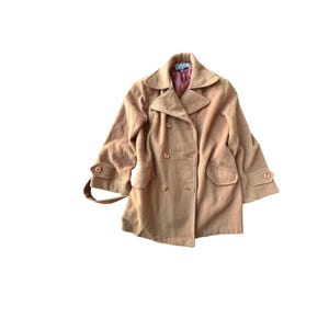 May include: A tan double-breasted coat featuring a wide collar, long sleeves with buttoned cuffs, and two front flap pockets. The coat has a visible label at the neckline and a dark pink interior lining visible at the collar opening.