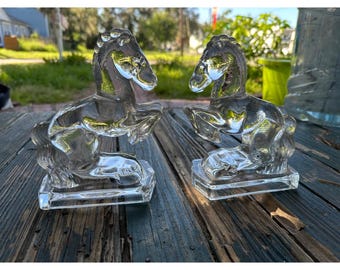 May include: A pair of clear glass horse figurines, each depicted in a rearing pose with front legs lifted. The horses are crafted from transparent, moulded glass and are mounted on rectangular glass bases, showcasing detailed manes and tails.