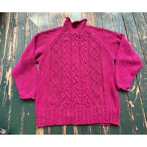 May include: A vibrant magenta long-sleeve sweater featuring a high turtleneck collar and a prominent central cable knit pattern. The garment displays vertical braided textures flanked by diamond-shaped knit designs, finished with a ribbed hem.