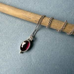 May include: A silver necklace with a deep red oval gemstone pendant. The pendant is set in a silver bezel and has a small silver bead at the bottom. The chain is delicate and silver, resting on a wooden dowel against a blue background.