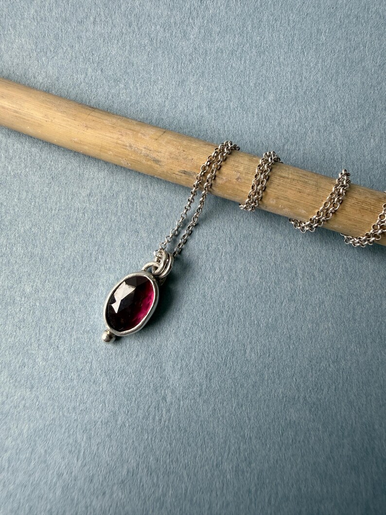 May include: A silver necklace with a deep red oval gemstone pendant. The pendant is set in a silver bezel and has a small silver bead at the bottom. The chain is delicate and silver, resting on a wooden dowel against a blue background.