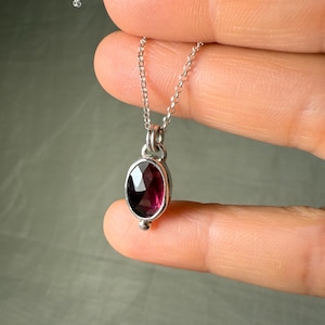 May include: A silver necklace with a small, oval, dark red gemstone pendant. The pendant is set in a silver bezel and has a small silver bead at the bottom. The chain is delicate and silver.