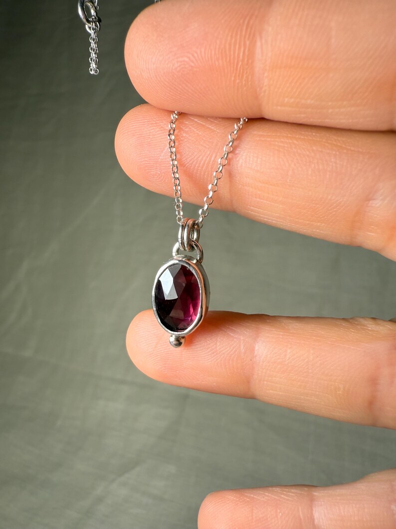 May include: A silver necklace with a small, oval, dark red gemstone pendant. The pendant is set in a silver bezel and has a small silver bead at the bottom. The chain is delicate and silver.