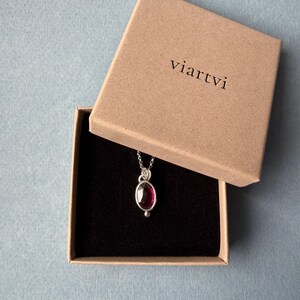 May include: A silver necklace with a dark red oval gemstone pendant, presented in an open brown box with the word "viartvi" printed on the lid. The box has a black interior, set against a light blue background.