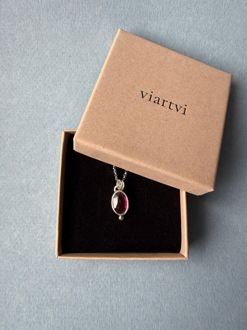 May include: A silver necklace with a dark red oval gemstone pendant, presented in an open brown box with the word "viartvi" printed on the lid. The box has a black interior, set against a light blue background.