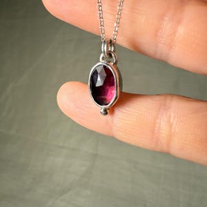 May include: A silver necklace featuring an oval, faceted, deep red gemstone pendant. The pendant is set in a silver bezel and hangs from a delicate silver chain. A small silver bead accents the bottom of the pendant.