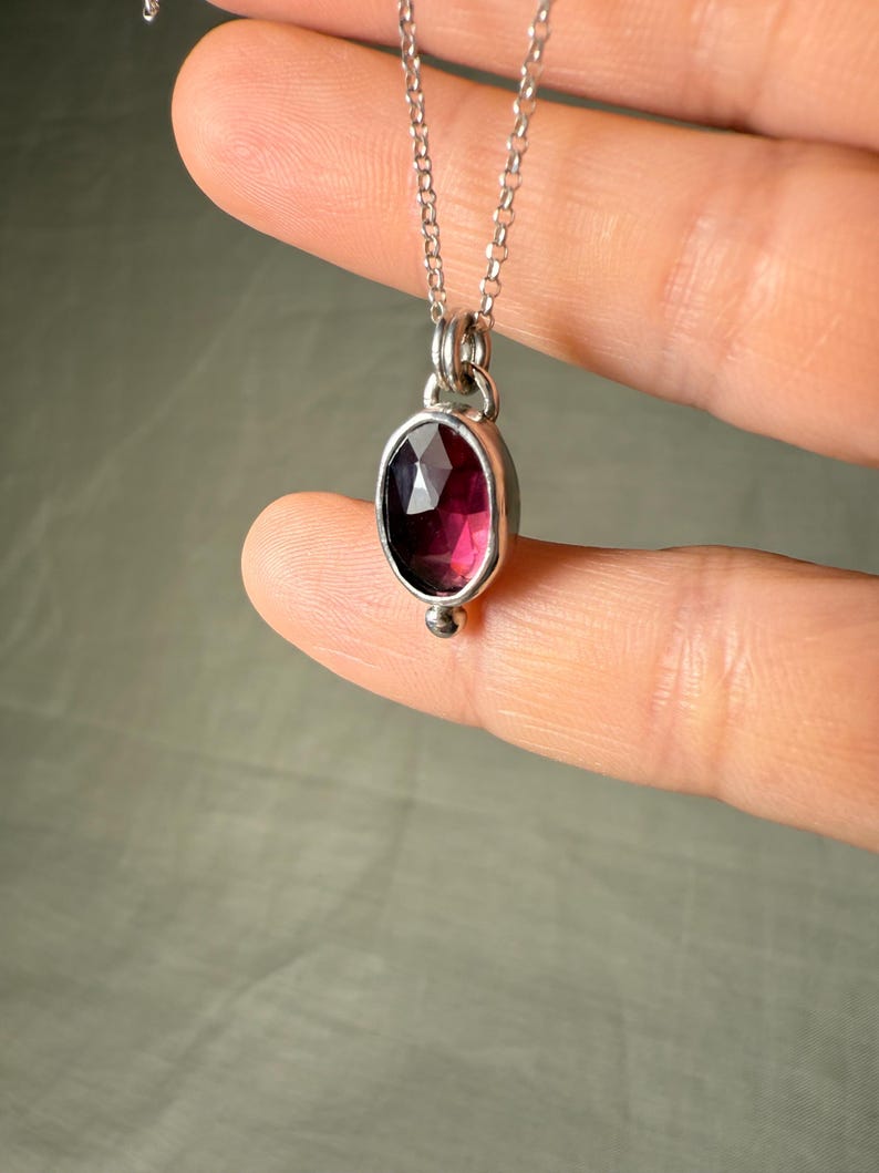 May include: A silver necklace featuring an oval, faceted, deep red gemstone pendant. The pendant is set in a silver bezel and hangs from a delicate silver chain. A small silver bead accents the bottom of the pendant.