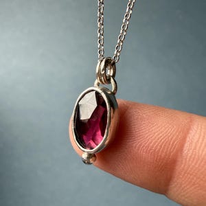 May include: A silver pendant necklace featuring a faceted, oval-shaped, deep red gemstone. The gemstone is set in a silver bezel and hangs from a delicate silver chain. A small silver bead accents the bottom of the pendant.