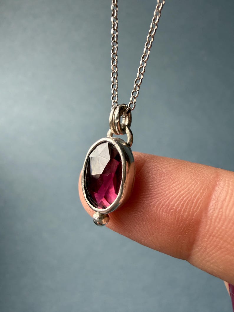 May include: A silver pendant necklace featuring a faceted, oval-shaped, deep red gemstone. The gemstone is set in a silver bezel and hangs from a delicate silver chain. A small silver bead accents the bottom of the pendant.