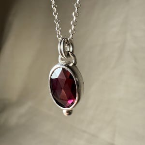 May include: A silver necklace featuring a pendant with a faceted, oval-shaped, deep red gemstone. The pendant is set in a silver bezel and hangs from a delicate silver chain. The gemstone has a rich, dark color, and the necklace is displayed against a neutral background.